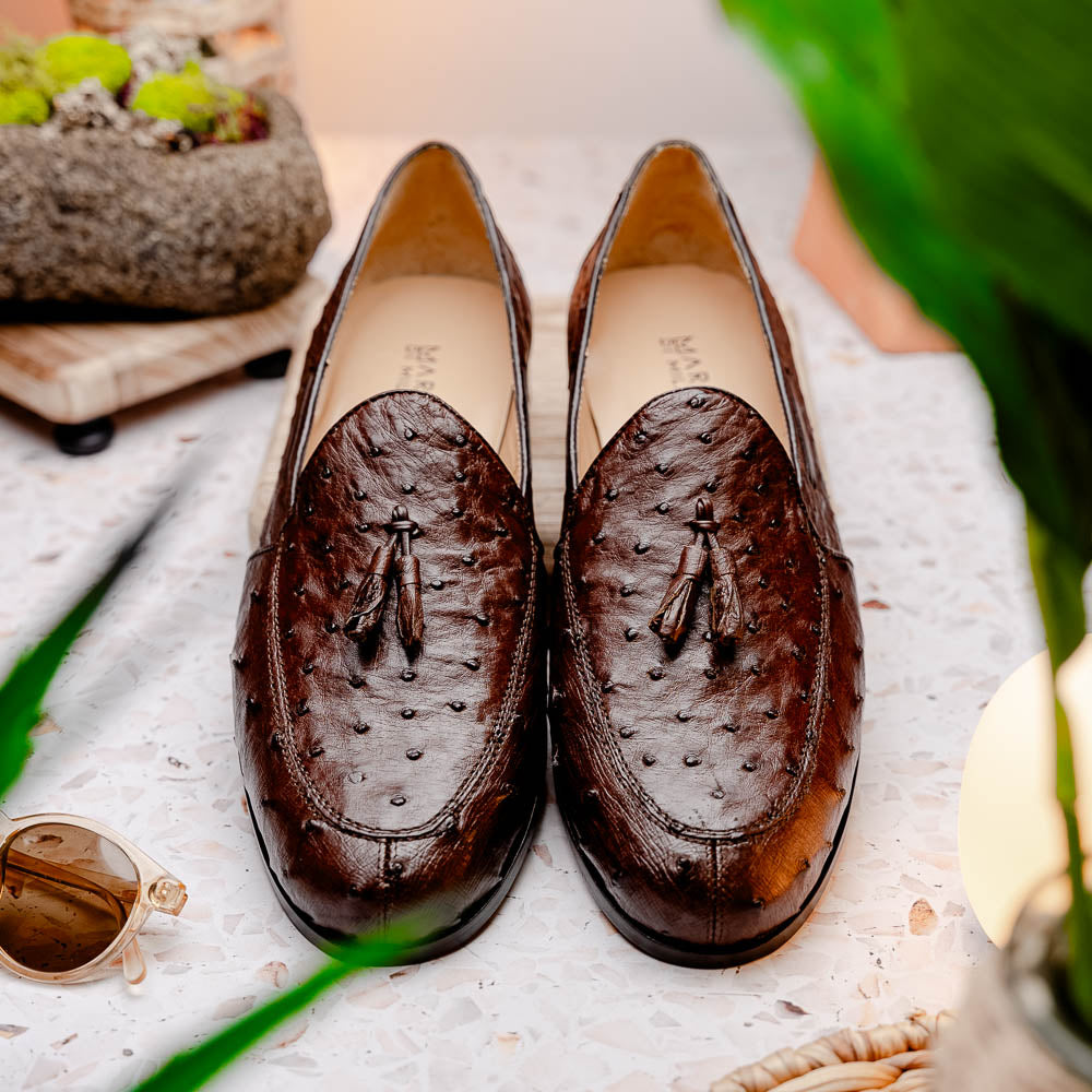 A pair of Marco Di Milano Aubiere Brown men's loafers, featuring brown leather with a textured pattern and tassel detail; one shoe stands upright while the other lies on its side showing the branded sole.