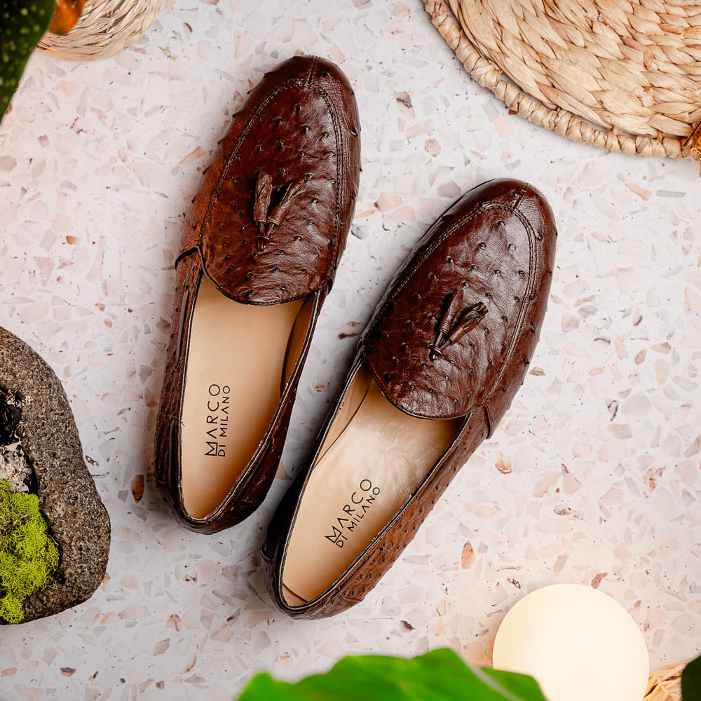 A pair of Marco Di Milano Aubiere Brown men's loafers, featuring brown leather with a textured pattern and tassel detail; one shoe stands upright while the other lies on its side showing the branded sole.
