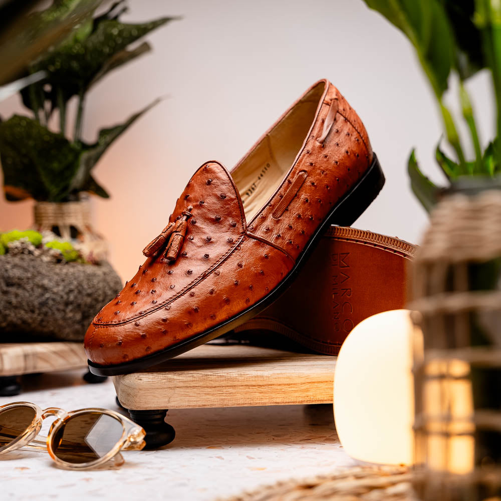A pair of luxury men's footwear—Aubiere Cognac loafers by Marco Di Milano—boasts rich brown ostrich leather, with one shoe upright and the other on its side to reveal a sole stamped "Marco Di Milano.