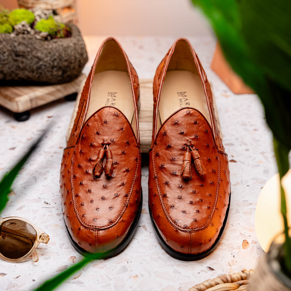 A pair of luxury men's footwear—Aubiere Cognac loafers by Marco Di Milano—boasts rich brown ostrich leather, with one shoe upright and the other on its side to reveal a sole stamped "Marco Di Milano.