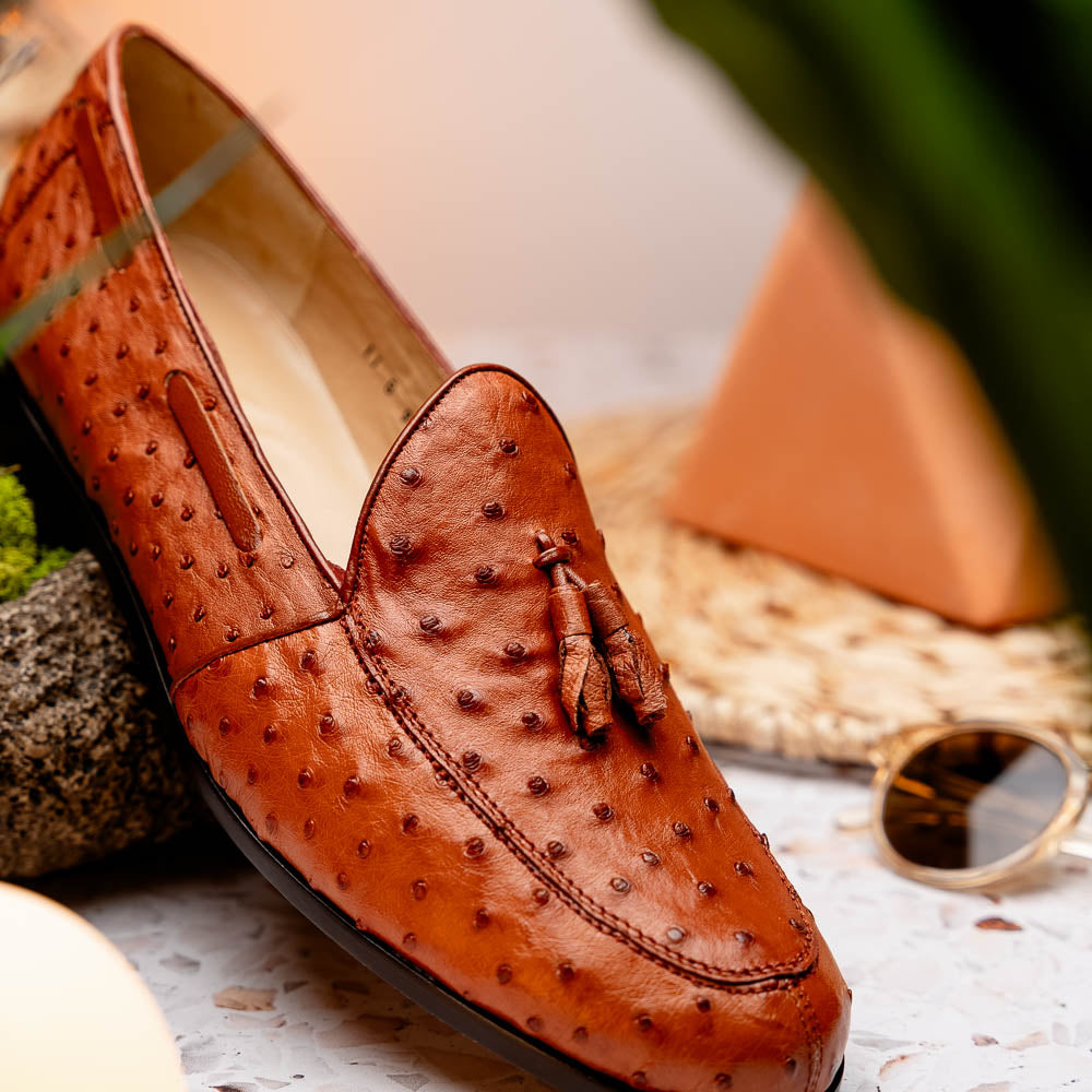 A pair of luxury men's footwear—Aubiere Cognac loafers by Marco Di Milano—boasts rich brown ostrich leather, with one shoe upright and the other on its side to reveal a sole stamped "Marco Di Milano.
