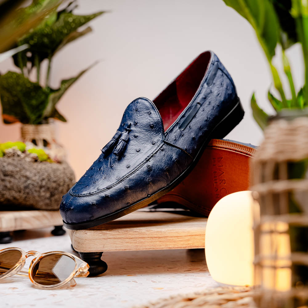 A pair of luxury men's footwear: the Aubiere Navy by Marco Di Milano—blue ostrich tassel loafers with red inner lining and brown leather soles, one shoe propped to display the sole branding.