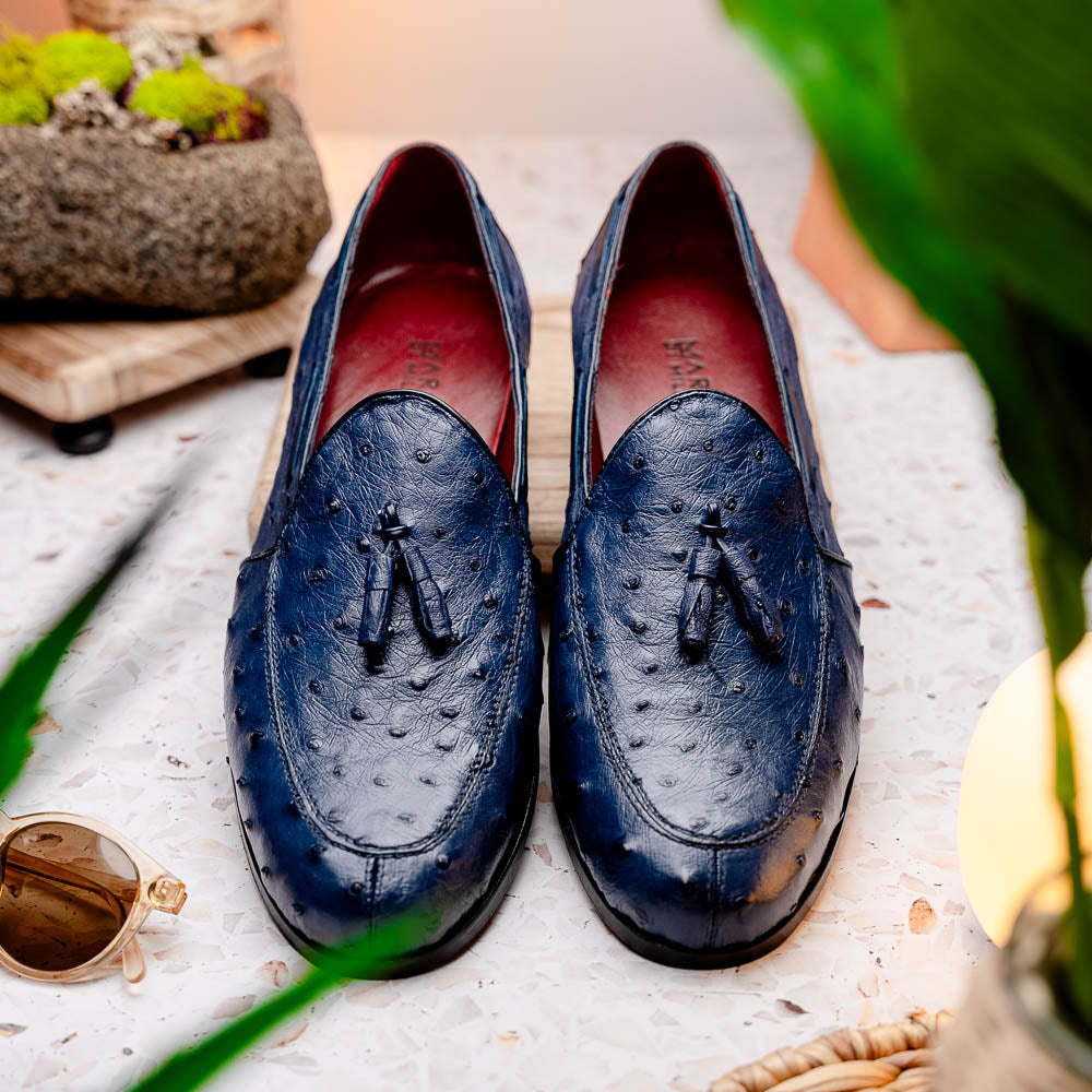 A pair of luxury men's footwear: the Aubiere Navy by Marco Di Milano—blue ostrich tassel loafers with red inner lining and brown leather soles, one shoe propped to display the sole branding.