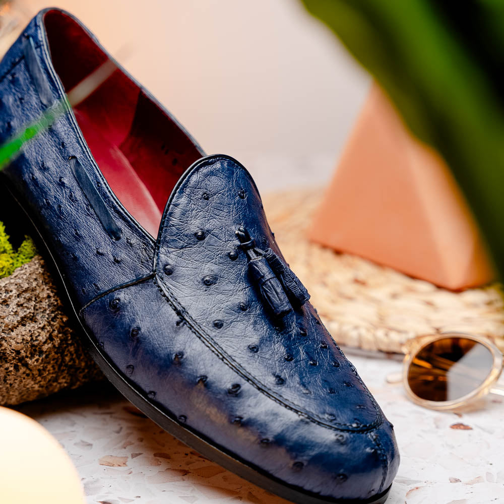 A pair of luxury men's footwear: the Aubiere Navy by Marco Di Milano—blue ostrich tassel loafers with red inner lining and brown leather soles, one shoe propped to display the sole branding.