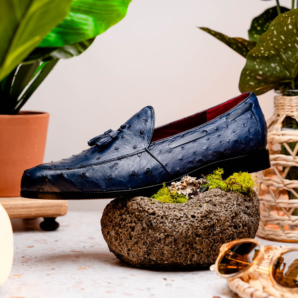 A pair of luxury men's footwear: the Aubiere Navy by Marco Di Milano—blue ostrich tassel loafers with red inner lining and brown leather soles, one shoe propped to display the sole branding.