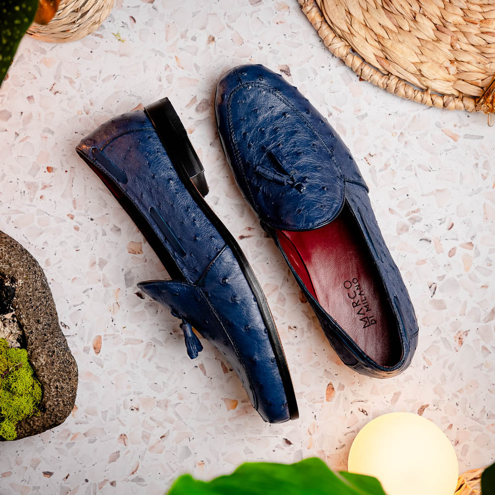 A pair of luxury men's footwear: the Aubiere Navy by Marco Di Milano—blue ostrich tassel loafers with red inner lining and brown leather soles, one shoe propped to display the sole branding.