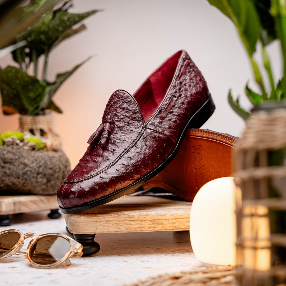 A pair of luxury Marco Di Milano Aubiere Wine loafers in burgundy ostrich leather—one upright showing tassels and quill detail, the other flipped to display the tan leather sole stamped “Marco Di Milano.”.