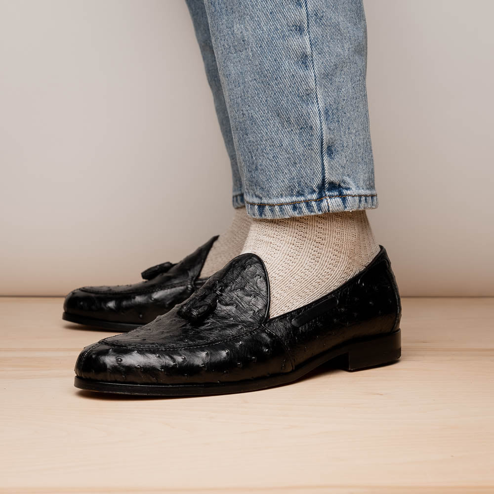 The Aubiere Black by Marco Di Milano are luxury men's tassel loafers in black genuine ostrich leather, shown with one shoe upright and the other displaying a tan sole embossed with the Marco Di Milano logo.