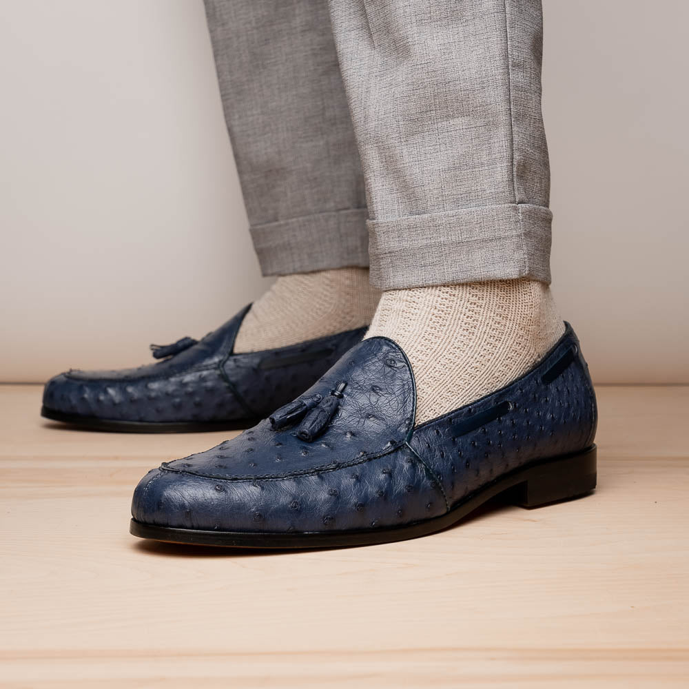 A pair of luxury men's footwear: the Aubiere Navy by Marco Di Milano—blue ostrich tassel loafers with red inner lining and brown leather soles, one shoe propped to display the sole branding.