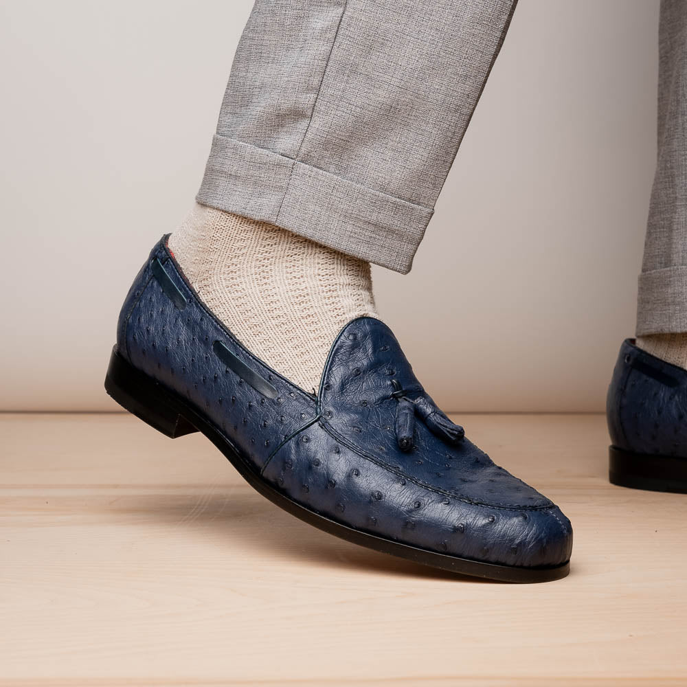 A pair of luxury men's footwear: the Aubiere Navy by Marco Di Milano—blue ostrich tassel loafers with red inner lining and brown leather soles, one shoe propped to display the sole branding.
