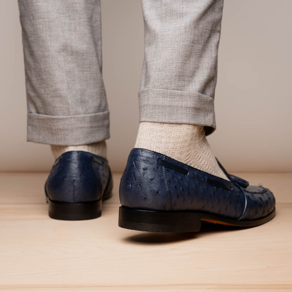 A pair of luxury men's footwear: the Aubiere Navy by Marco Di Milano—blue ostrich tassel loafers with red inner lining and brown leather soles, one shoe propped to display the sole branding.