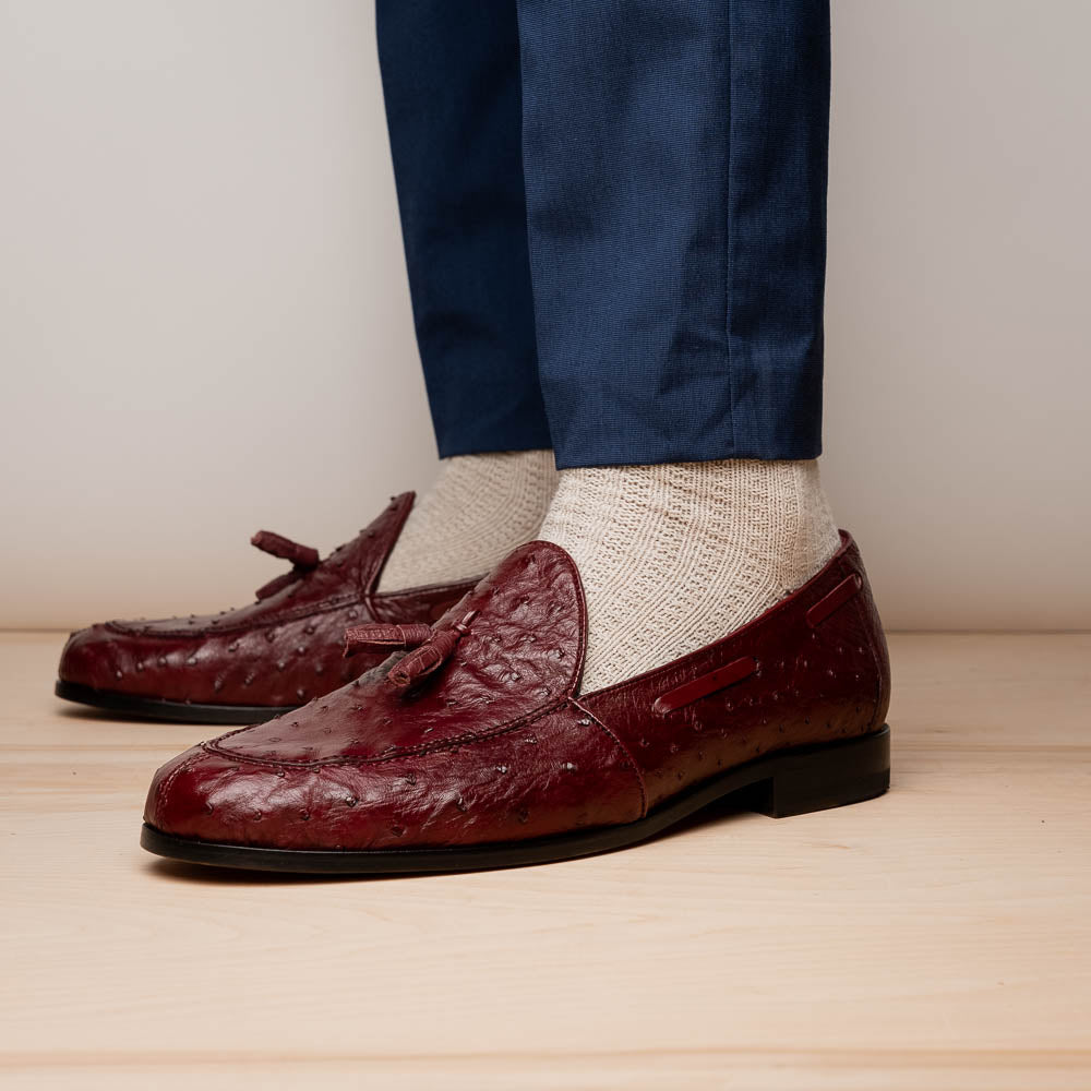 A pair of luxury Marco Di Milano Aubiere Wine loafers in burgundy ostrich leather—one upright showing tassels and quill detail, the other flipped to display the tan leather sole stamped “Marco Di Milano.”.