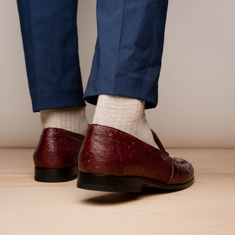 A pair of luxury Marco Di Milano Aubiere Wine loafers in burgundy ostrich leather—one upright showing tassels and quill detail, the other flipped to display the tan leather sole stamped “Marco Di Milano.”.