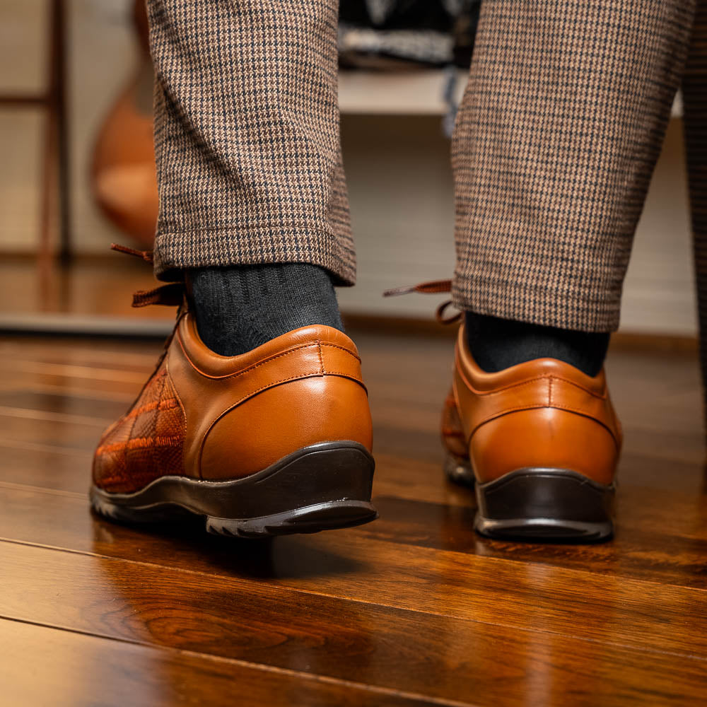 The Marco Di Milano Bari Cognac features brown leather with a hand-painted finish, textured side panels, black laces, and black rubber soles—one shoe propped against the other.