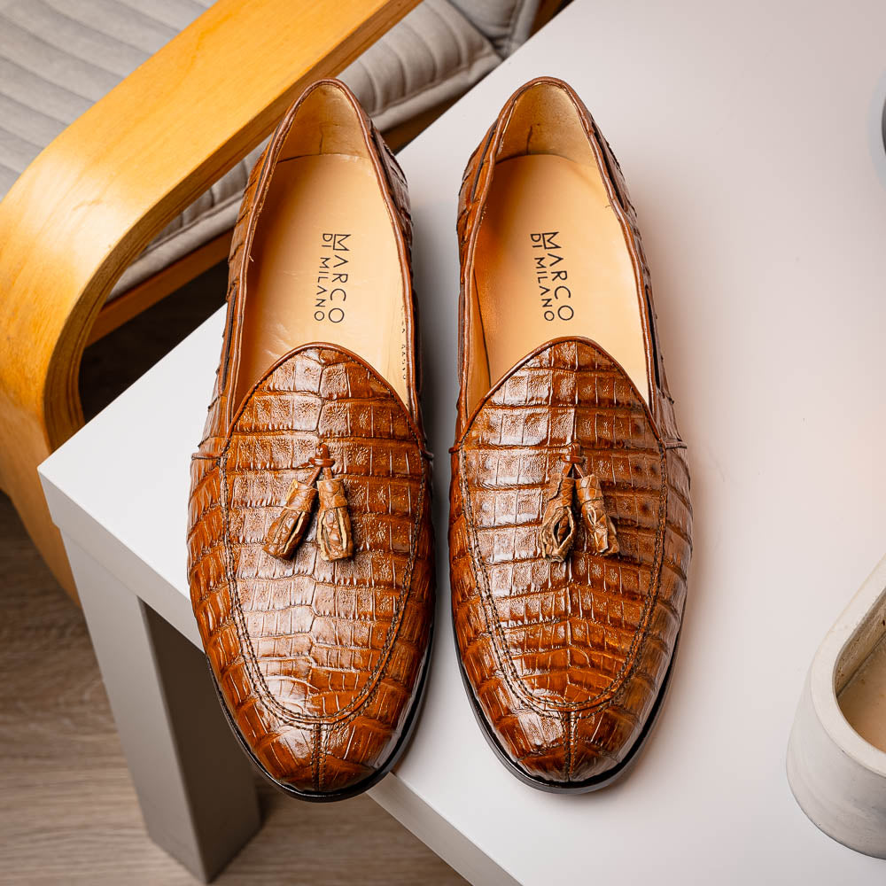 A person wearing dark blue dress pants and black socks with Marco Di Milano's Bianchi Bay Apache luxury tassel loafers—crafted from genuine caiman crocodile leather—stands on a white background.