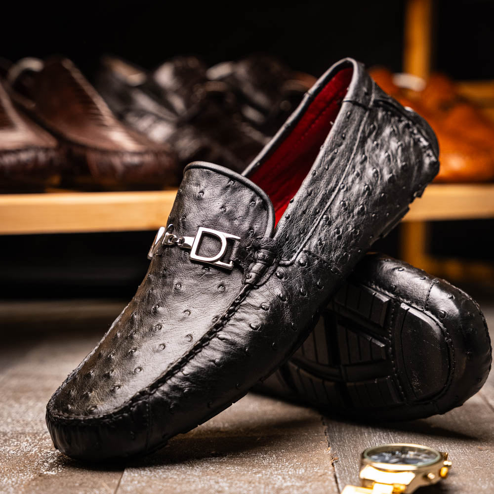 Bolonia Black by Marco Di Milano: a pair of black leather loafers with textured finish and metal buckle, one shoe upright and the other propped against it, displayed on a white background.