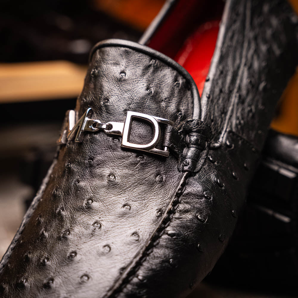 Bolonia Black by Marco Di Milano: a pair of black leather loafers with textured finish and metal buckle, one shoe upright and the other propped against it, displayed on a white background.