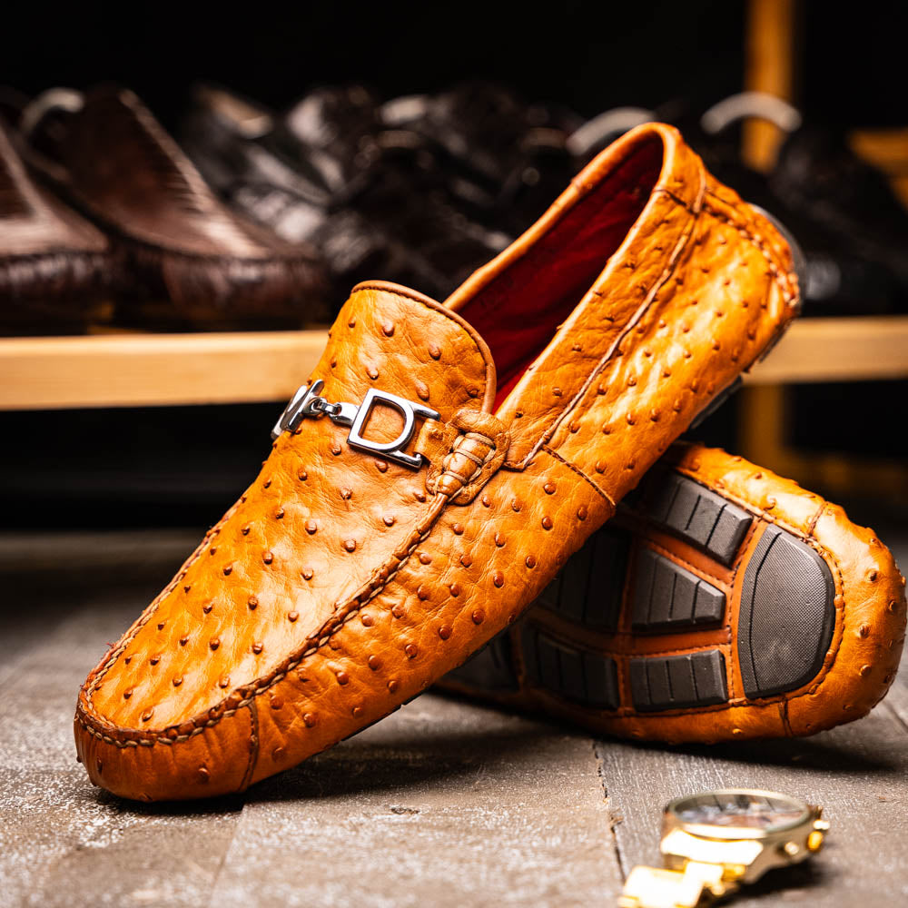 The Bolonia Brandy by Marco Di Milano—brown ostrich leather loafers with hand-painted finishes and metal buckles, one upright and one on its side revealing the sole—exude luxury, crafted from genuine exotic leather.