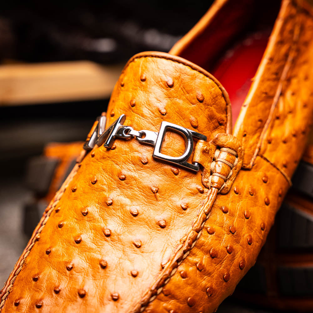 The Bolonia Brandy by Marco Di Milano—brown ostrich leather loafers with hand-painted finishes and metal buckles, one upright and one on its side revealing the sole—exude luxury, crafted from genuine exotic leather.