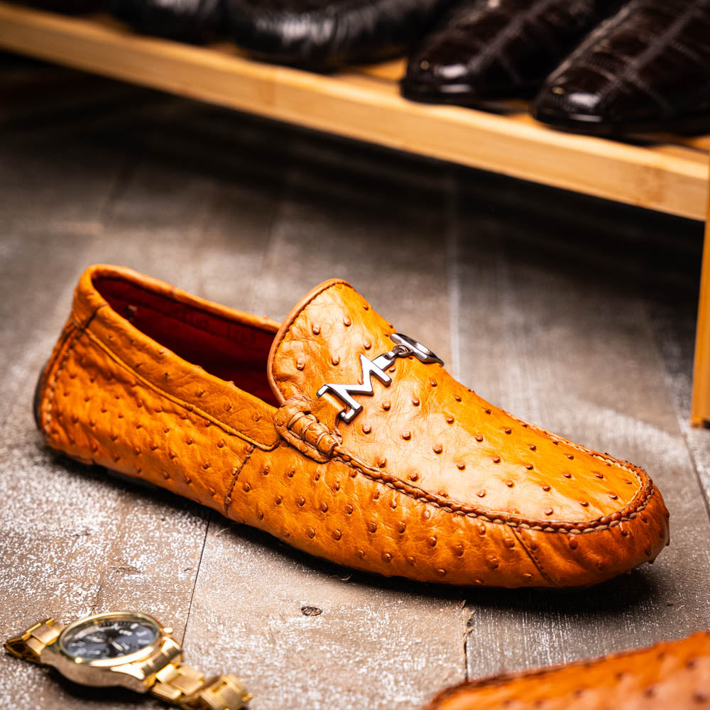 The Bolonia Brandy by Marco Di Milano—brown ostrich leather loafers with hand-painted finishes and metal buckles, one upright and one on its side revealing the sole—exude luxury, crafted from genuine exotic leather.