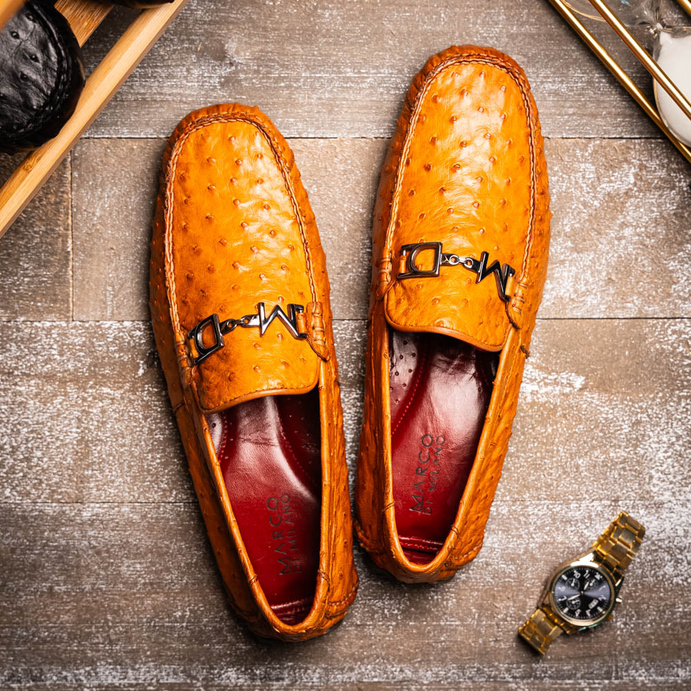 The Bolonia Brandy by Marco Di Milano—brown ostrich leather loafers with hand-painted finishes and metal buckles, one upright and one on its side revealing the sole—exude luxury, crafted from genuine exotic leather.