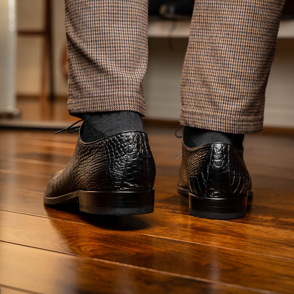 The Marco Di Milano Bono Black features a pair of luxury dress shoes in a crocodile pattern—one upright and one displaying a brown branded sole.