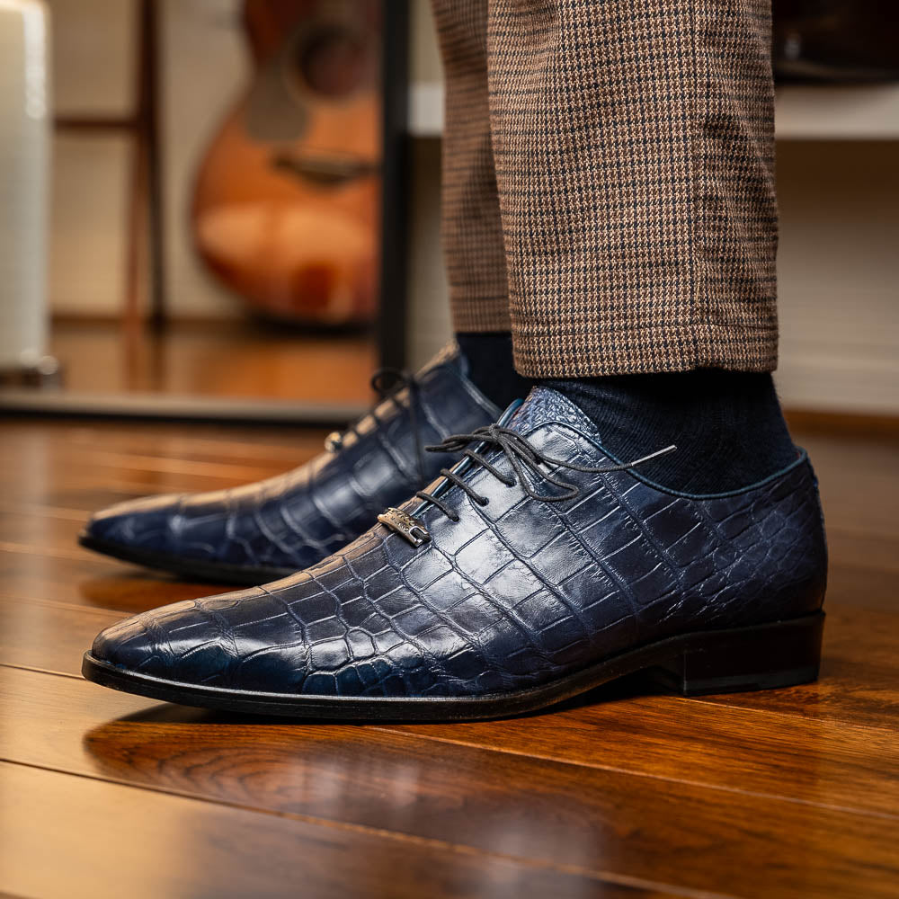 The Bono Navy by Marco Di Milano: luxury navy blue dress shoes with crocodile-patterned leather, black soles, and red inner lining. One shoe is positioned to showcase the sole.