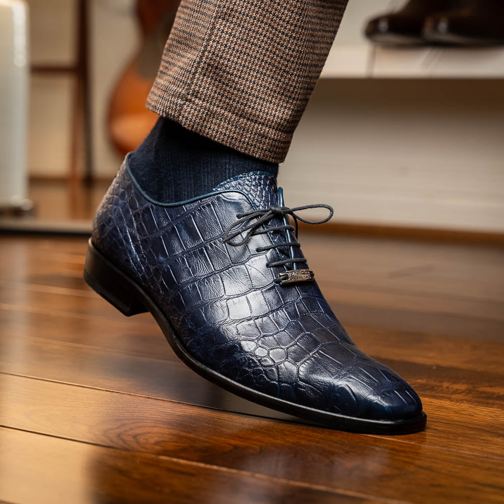 The Bono Navy by Marco Di Milano: luxury navy blue dress shoes with crocodile-patterned leather, black soles, and red inner lining. One shoe is positioned to showcase the sole.