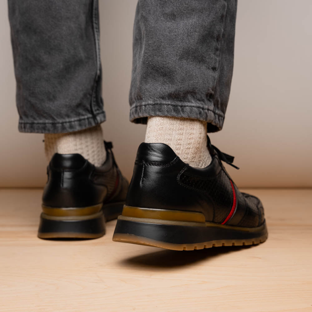The Marco Di Milano Brescia Black sneakers feature textured black leather, black laces, a red side stripe, and brown rubber soles—one shoe stands upright while the other lies on its side.