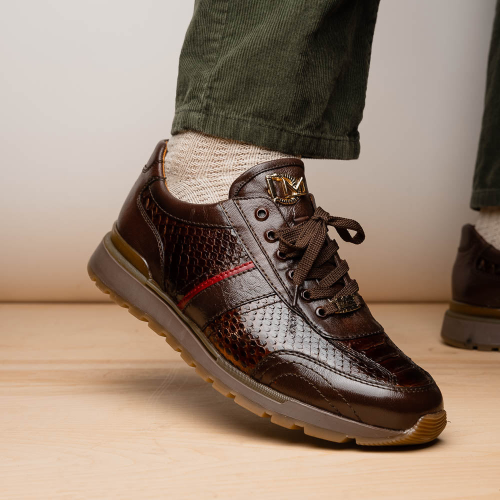 The Marco Di Milano Brescia Brown sneakers feature dark brown python-textured leather, a gold logo on the tongue, and a red stripe detail, with one shoe propped on the other—ideal for luxury footwear enthusiasts.