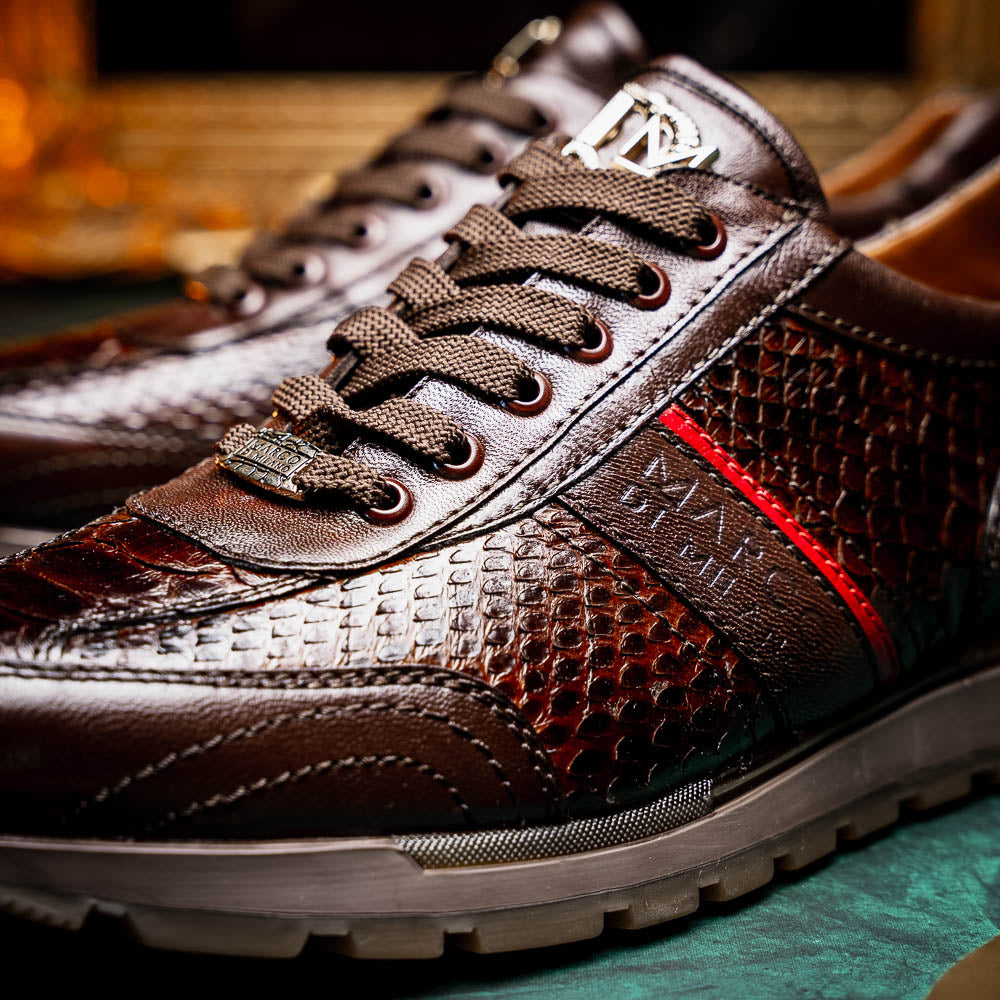 The Marco Di Milano Brescia Brown sneakers feature dark brown python-textured leather, a gold logo on the tongue, and a red stripe detail, with one shoe propped on the other—ideal for luxury footwear enthusiasts.