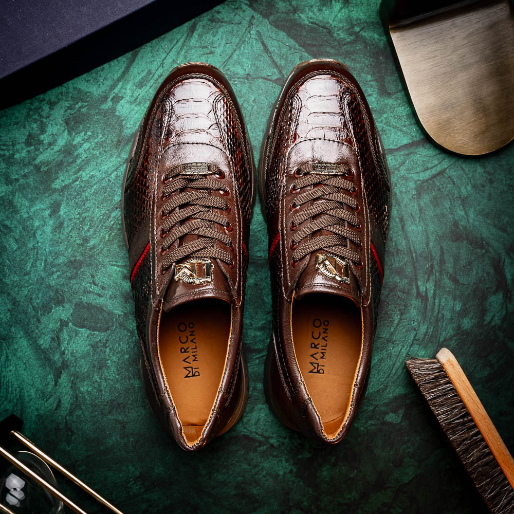 The Marco Di Milano Brescia Brown sneakers feature dark brown python-textured leather, a gold logo on the tongue, and a red stripe detail, with one shoe propped on the other—ideal for luxury footwear enthusiasts.