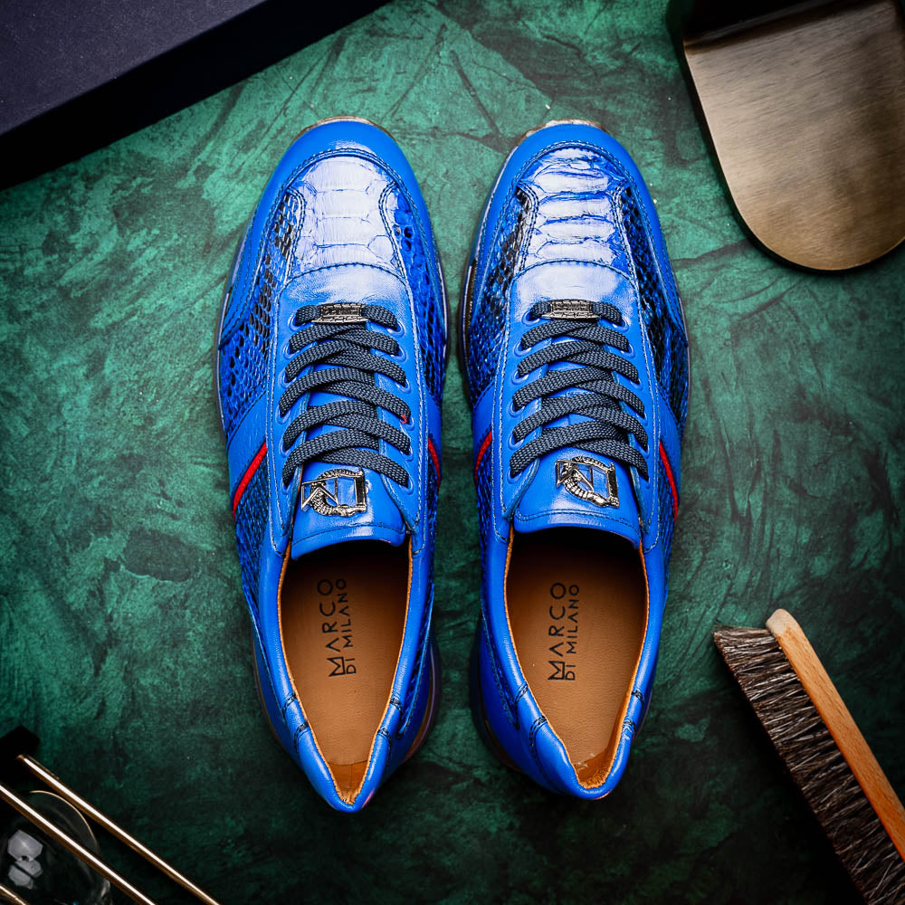 The Brescia Electric Blue sneakers by Marco Di Milano feature blue python leather, black laces, a red stripe, hand-painted side panels, and brown rubber soles—displayed with one shoe upright and the other on its side.