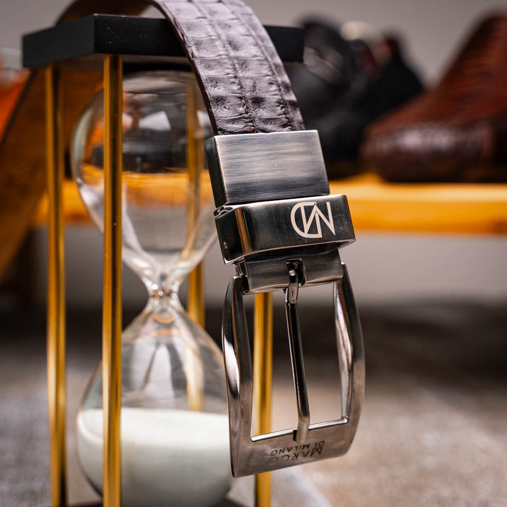 The Marco Di Milano Caribe Belt Brown / Pantano, a luxurious men's belt crafted from exotic caiman fuscus leather in brown and black with a silver buckle, is coiled against a white background.