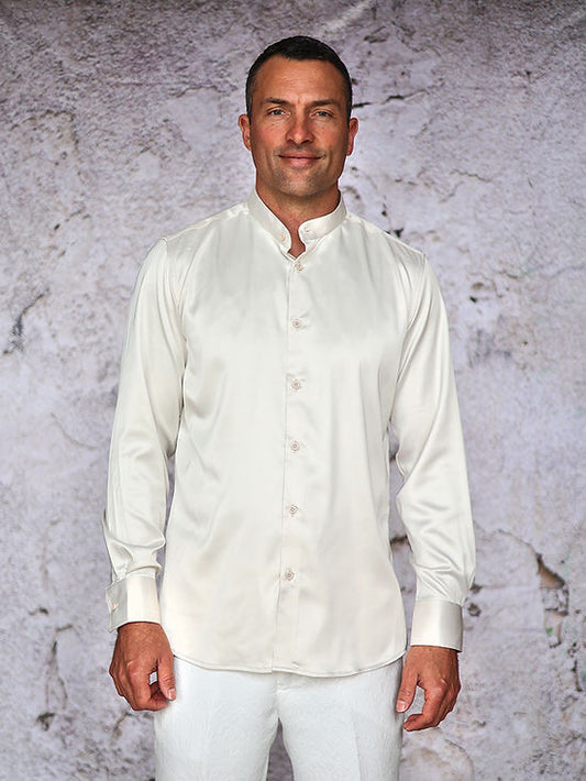 A man stands against a textured white wall, wearing the Manzini MANZINI INR-102 Banded Collar Solid Color Satin Cream Shirt and white pants.