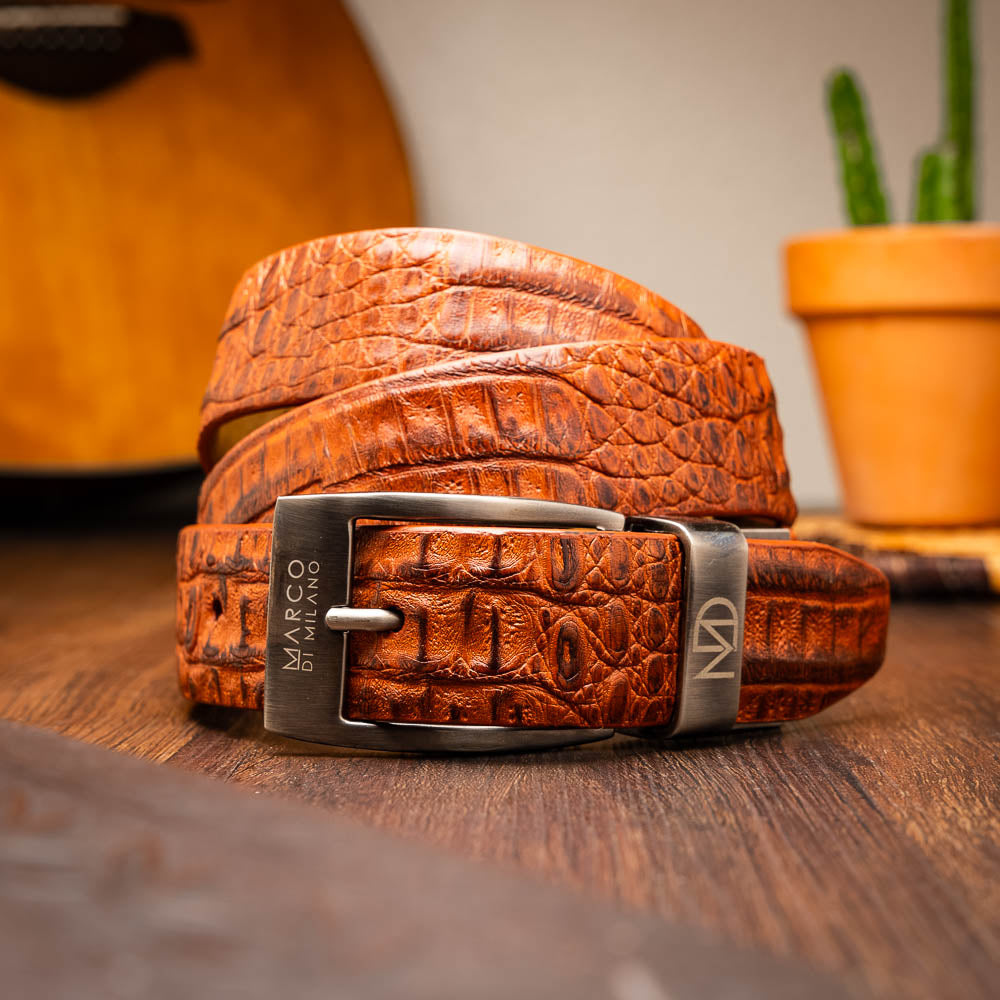The Riccardo Belt Brandy by Marco Di Milano is a brown exotic leather belt with a crocodile pattern, silver buckle engraved with "Marco Di Milano" and "MD," styled after caiman fuscus-belly belts for a refined look.