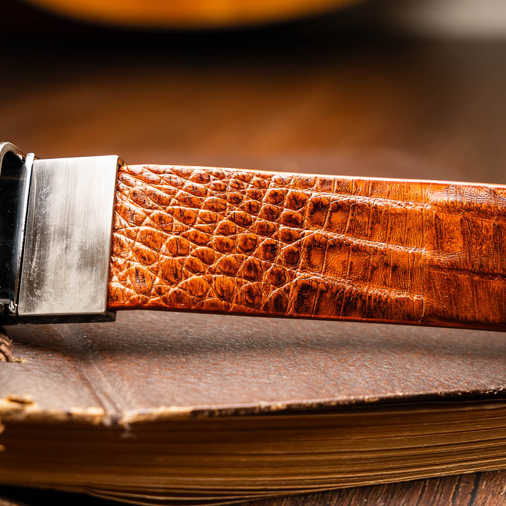 The Riccardo Belt Brandy by Marco Di Milano is a brown exotic leather belt with a crocodile pattern, silver buckle engraved with "Marco Di Milano" and "MD," styled after caiman fuscus-belly belts for a refined look.