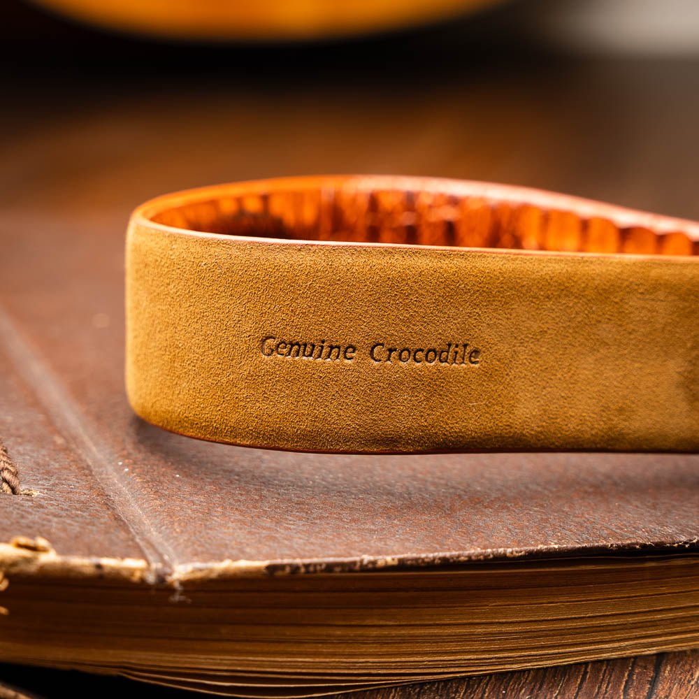 The Riccardo Belt Brandy by Marco Di Milano is a brown exotic leather belt with a crocodile pattern, silver buckle engraved with "Marco Di Milano" and "MD," styled after caiman fuscus-belly belts for a refined look.