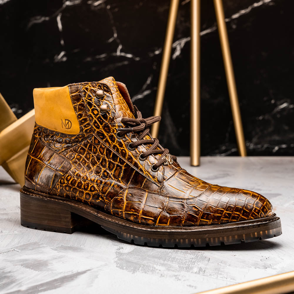 The Potenza Mustard/Brown boots by Marco Di Milano feature brown crocodile-pattern leather, lace-up fronts, tan ankle detail, and textured rubber soles with hand-painted finishes—one boot upright, the other on its side.