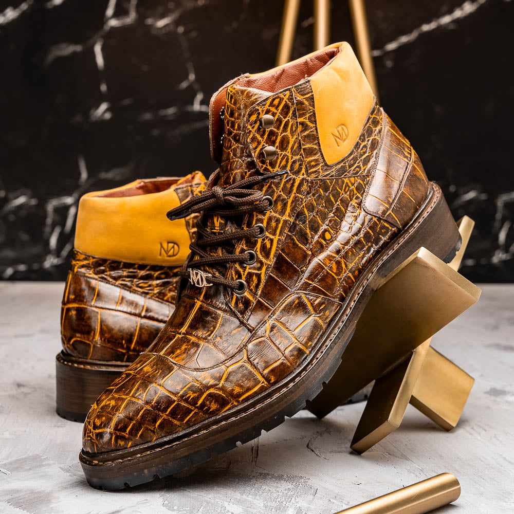 The Potenza Mustard/Brown boots by Marco Di Milano feature brown crocodile-pattern leather, lace-up fronts, tan ankle detail, and textured rubber soles with hand-painted finishes—one boot upright, the other on its side.