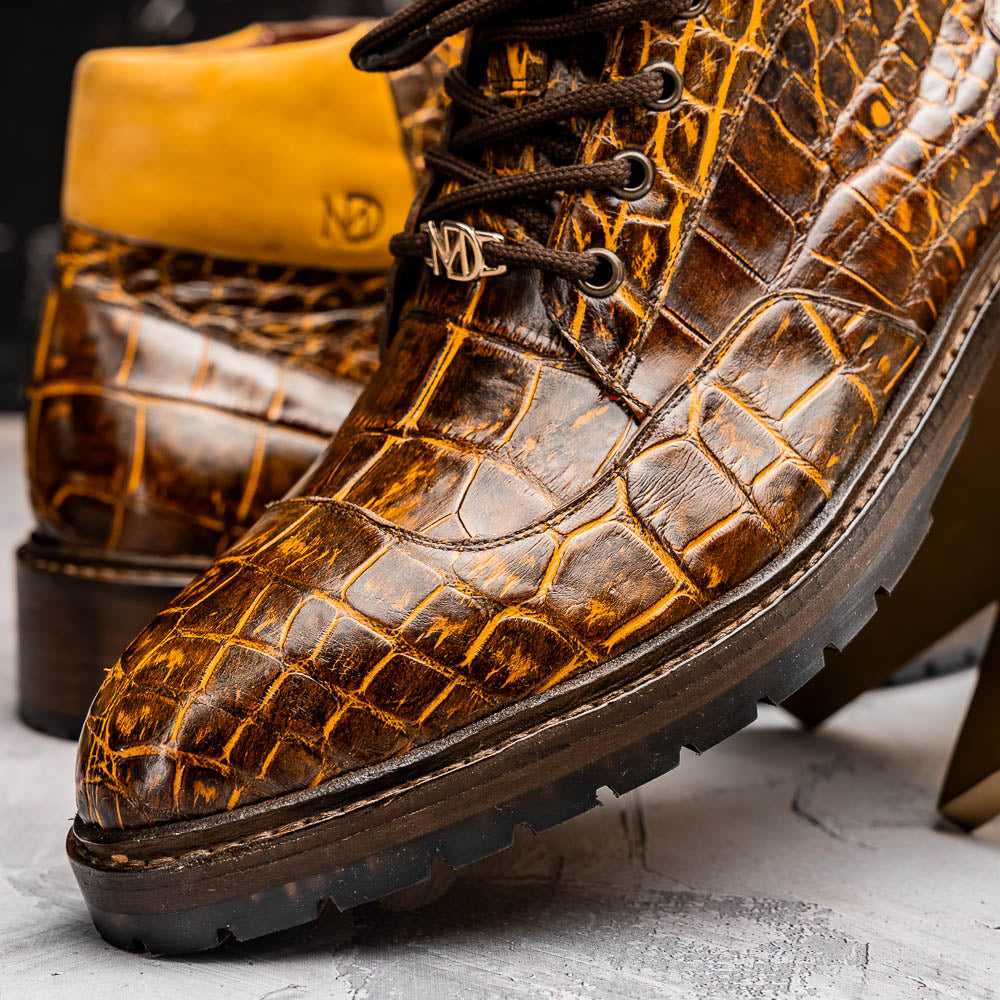 The Potenza Mustard/Brown boots by Marco Di Milano feature brown crocodile-pattern leather, lace-up fronts, tan ankle detail, and textured rubber soles with hand-painted finishes—one boot upright, the other on its side.