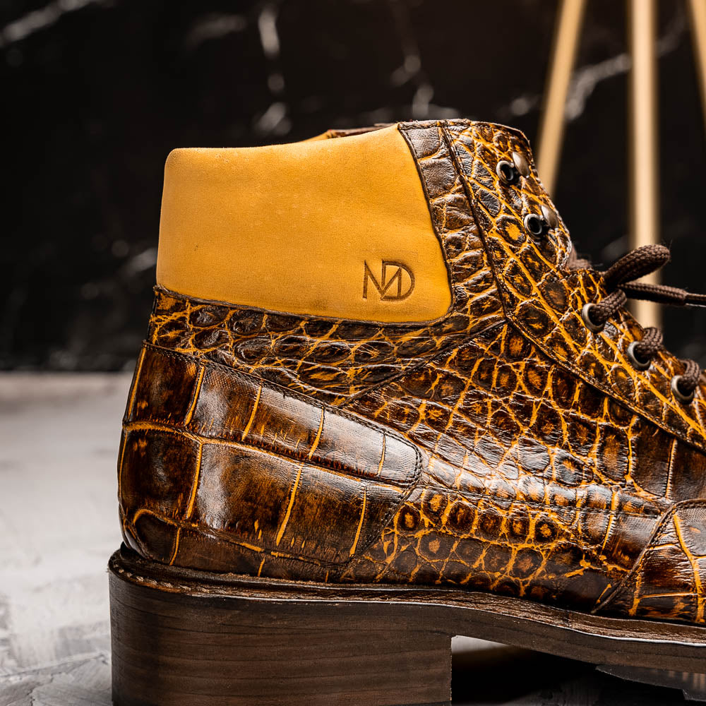 The Potenza Mustard/Brown boots by Marco Di Milano feature brown crocodile-pattern leather, lace-up fronts, tan ankle detail, and textured rubber soles with hand-painted finishes—one boot upright, the other on its side.