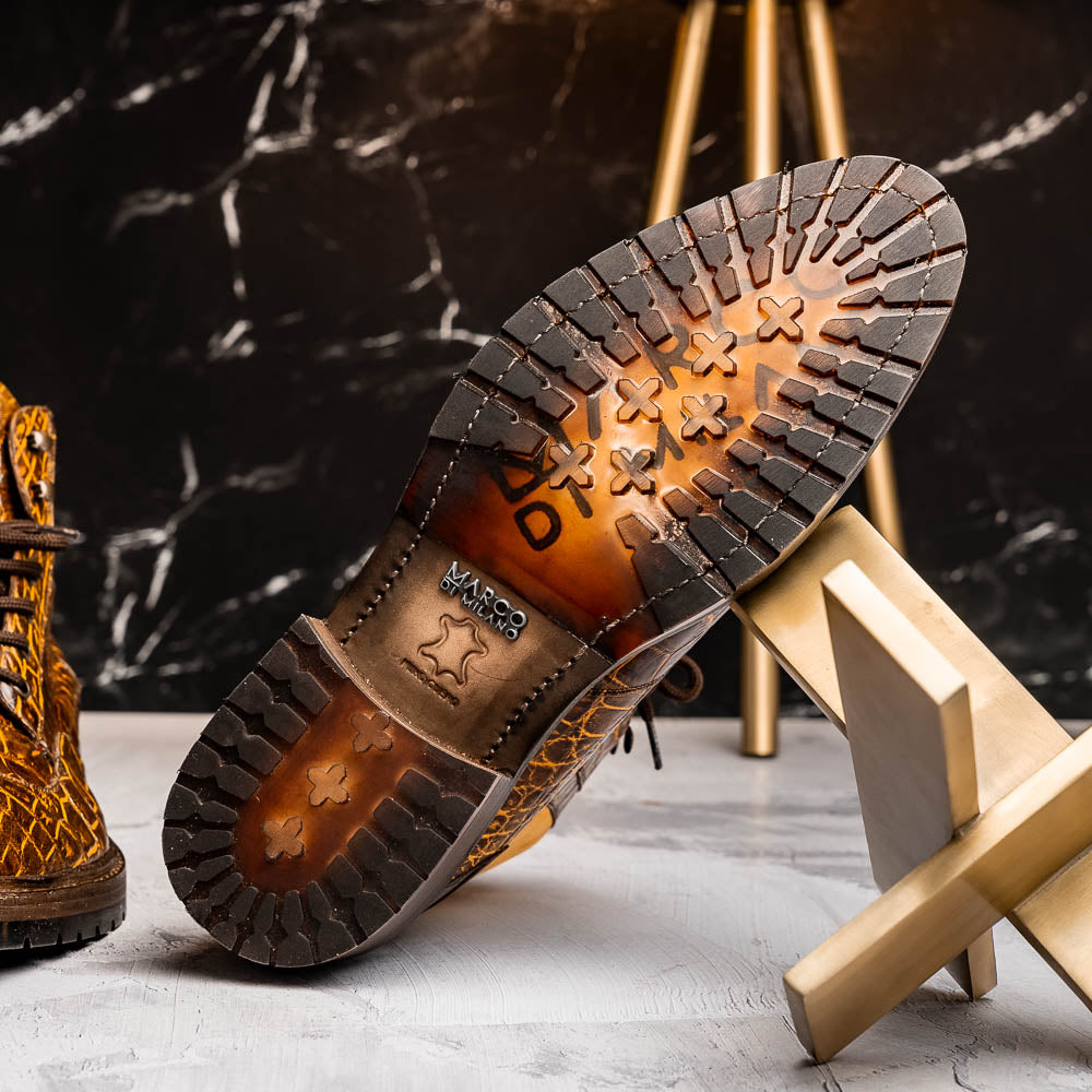 The Potenza Mustard/Brown boots by Marco Di Milano feature brown crocodile-pattern leather, lace-up fronts, tan ankle detail, and textured rubber soles with hand-painted finishes—one boot upright, the other on its side.