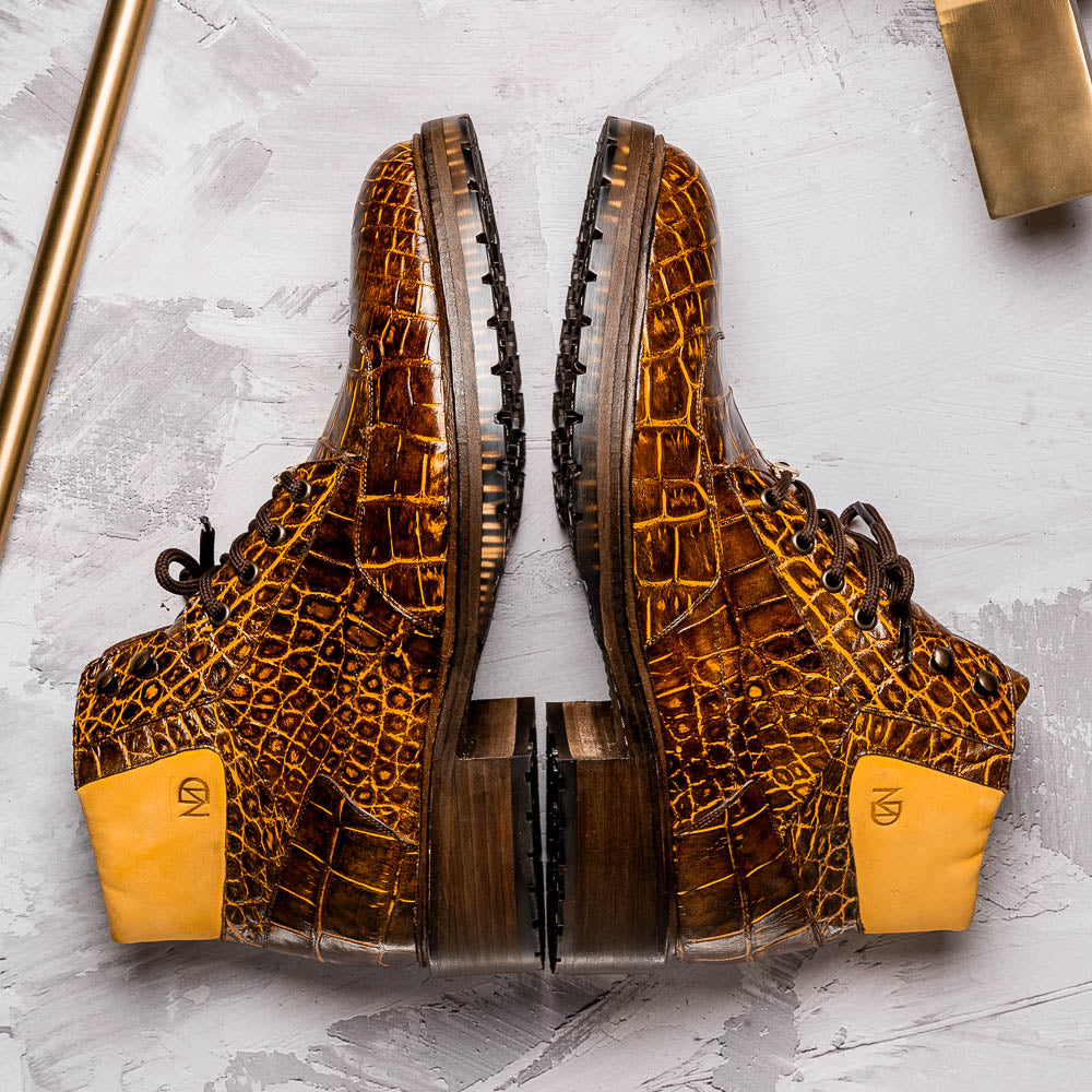 The Potenza Mustard/Brown boots by Marco Di Milano feature brown crocodile-pattern leather, lace-up fronts, tan ankle detail, and textured rubber soles with hand-painted finishes—one boot upright, the other on its side.
