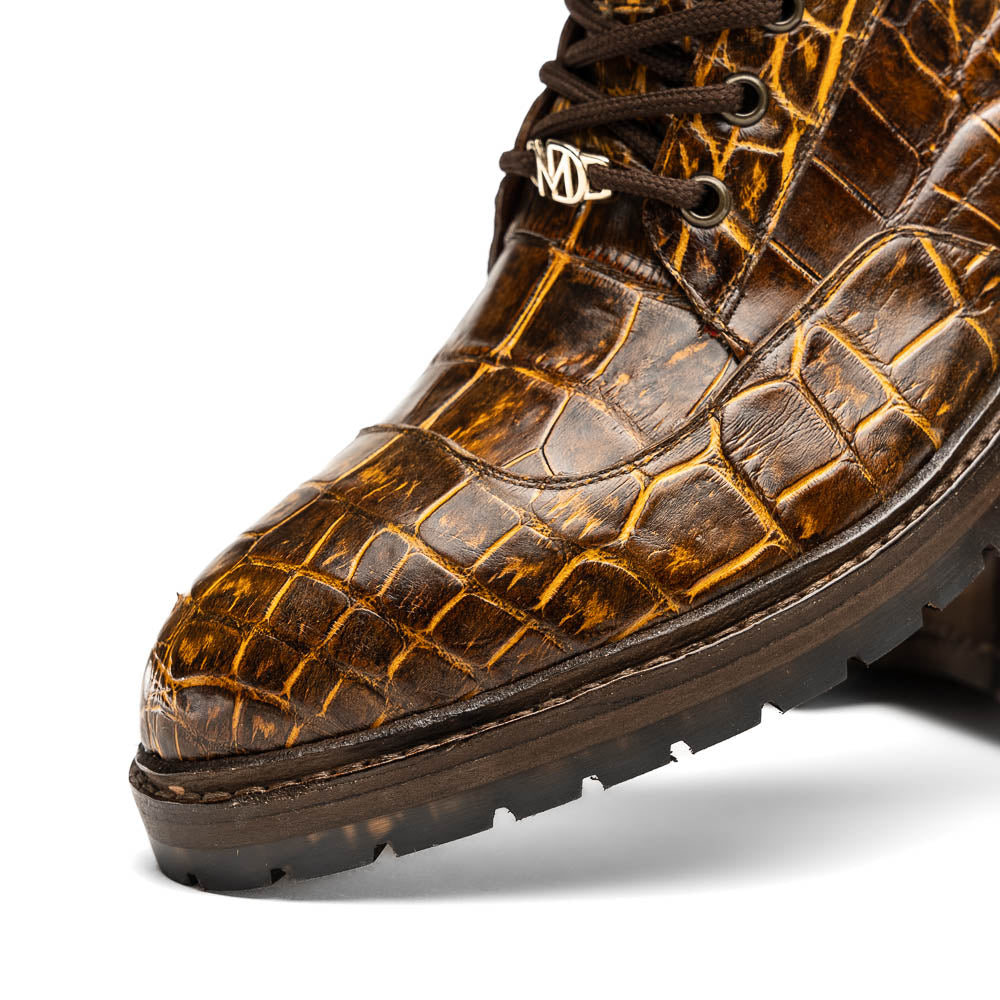 The Potenza Mustard/Brown boots by Marco Di Milano feature brown crocodile-pattern leather, lace-up fronts, tan ankle detail, and textured rubber soles with hand-painted finishes—one boot upright, the other on its side.