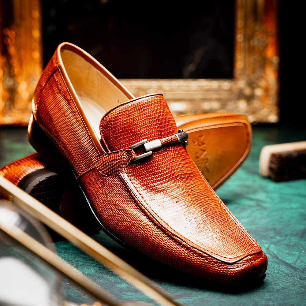 The Marco Di Milano Fabro-Liz Cognac loafers, crafted from brown leather with a textured finish and metal detail, are displayed—one upright and one showing the sole and branding—showcasing refined luxury footwear.