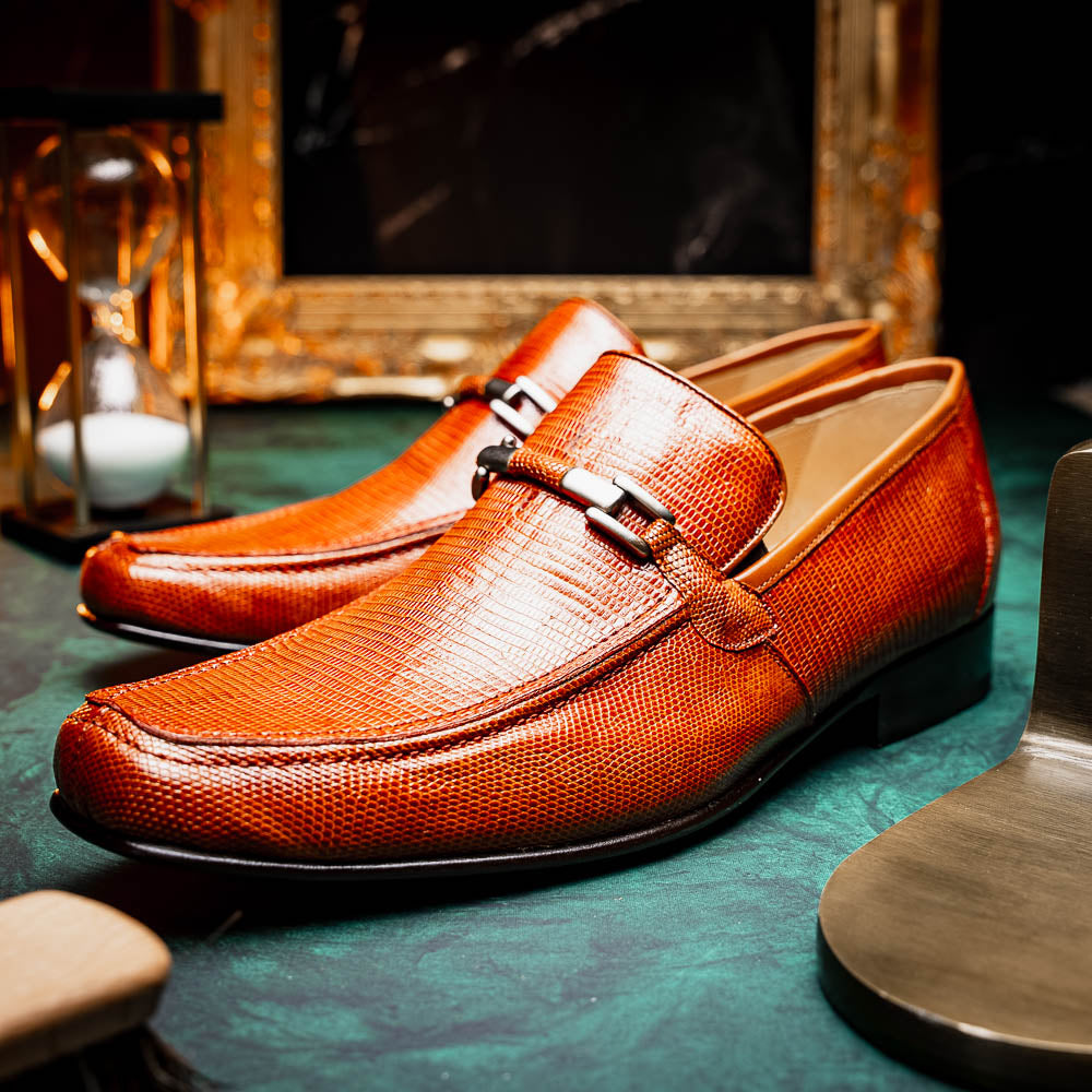 The Marco Di Milano Fabro-Liz Cognac loafers, crafted from brown leather with a textured finish and metal detail, are displayed—one upright and one showing the sole and branding—showcasing refined luxury footwear.