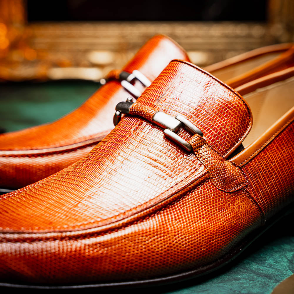 The Marco Di Milano Fabro-Liz Cognac loafers, crafted from brown leather with a textured finish and metal detail, are displayed—one upright and one showing the sole and branding—showcasing refined luxury footwear.