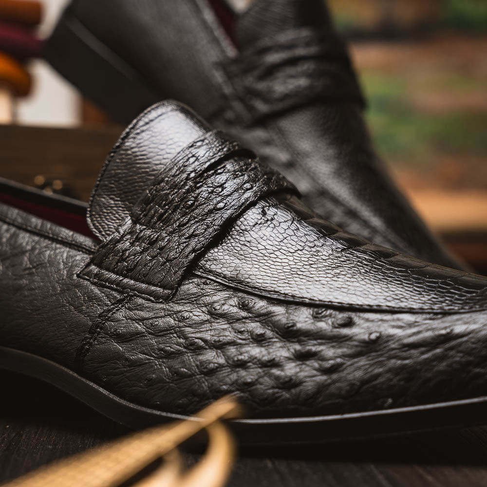 The Fangio Black by Marco Di Milano: a pair of black textured leather loafers, one upright and one sole-up, displaying the brand on the tan sole—luxury men's footwear to elevate any look.