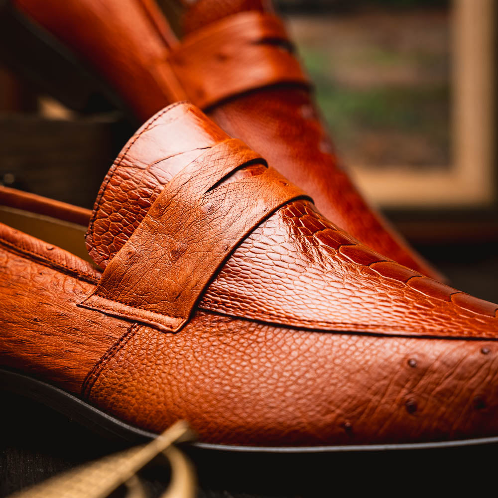 A pair of Marco Di Milano Fangio Cognac men's shoes in brown leather with textured detailing; one genuine ostrich loafer stands upright, while the other lies on its side showing the sole embossed with the brand name.